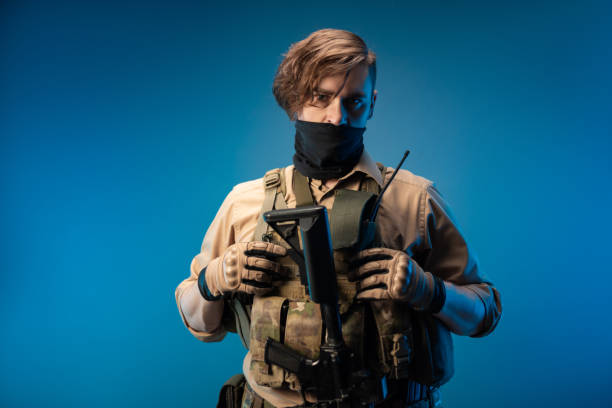 um soldado agente especial masculino em um colete à prova de balas com um rifle automático em um fundo azul - marines camouflage camouflage clothing male - fotografias e filmes do acervo