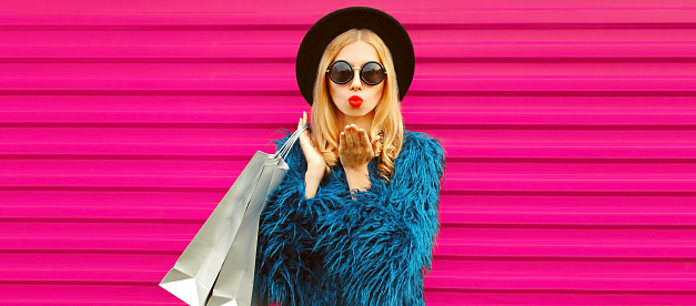 Portrait of beautiful stylish woman with shopping bags and blowing her lips sends air kiss with lipstick wearing blue fur coat, black round hat and sunglasses posing on pink background