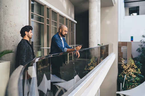 Side view of serious ethnic male in casual wear browsing smartphone talking to coworker discussing strategy of project leaning on fence at balcony