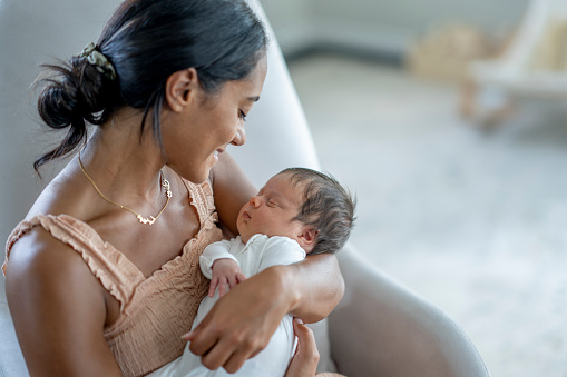 A new mother rocks her baby gently in her arms while sitting in the nursery and enjoying some one-on-one time bonding with him. She is dressed comfortably and the baby is wearing a sleeper as she gazes down at him with pride.