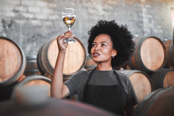 mujer negra degustando vino en una bodega, mirando y comprobando el color y la calidad de los años que produce. joven sommelier afroamericano orgulloso de la nueva incorporación, analizando vino blanco en una bodega - winemaking vintner winery people fotografías e imágenes de stock