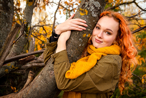 A young red haired woman hugs a tree trunk in an autumn park.