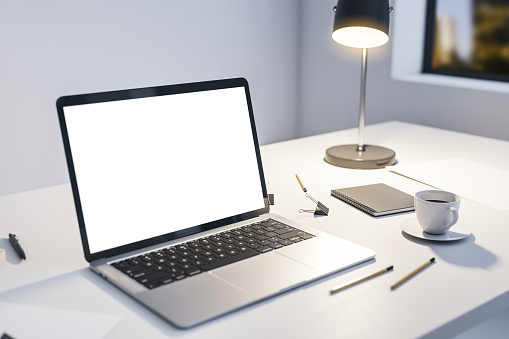 Perspective view on modern laptop blank white screen with empty space on light table with illuminated lamp, office tools and coffee cup and night city view from the window. 3D rendering, mock up
