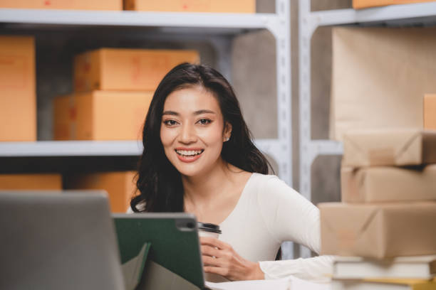 online shopping concept, happy young Asian business owner woman prepare parcel box and check orders of product for deliver to customer on laptop computer at home, e-business marketing on cyberspace concepto de compra en línea, joven y feliz mujer asiática propietaria de negocios preparar la caja de paquetes y verificar los pedidos de productos para entregarlos al cliente en una computadora portátil en casa, marketing de negocios electrónicos en e