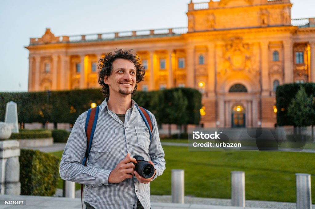 Young Tourist Enjoying The View Of The City Handsome young man enjoying the view in Stockholm, Sweden. Standing in front of a Parliament House (Riksdagshuset) building, holding a camera. Adult Stock Photo