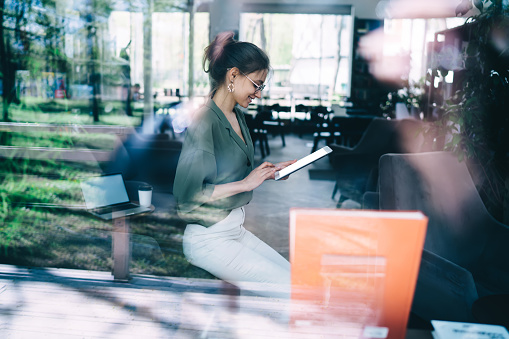 Side view through glass of modern pleased brunette female in glasses wearing casual clothes browsing tablet with joy while sitting in office