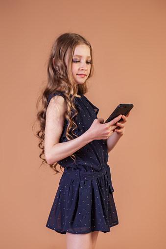 Portrait teenage girl 10 year old with curly long hair texting on mobile phone at beige background, talking and smiling. Fashion model in stylish blue dress. Fashionable young lady model. Copy space
