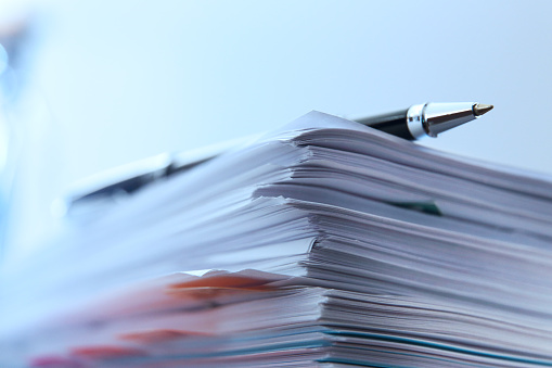 A ballpoint pen rests on top of a stack of documents ready for signing. The image is photographed using a very shallow depth of field with the focus being on the tip of the pen.