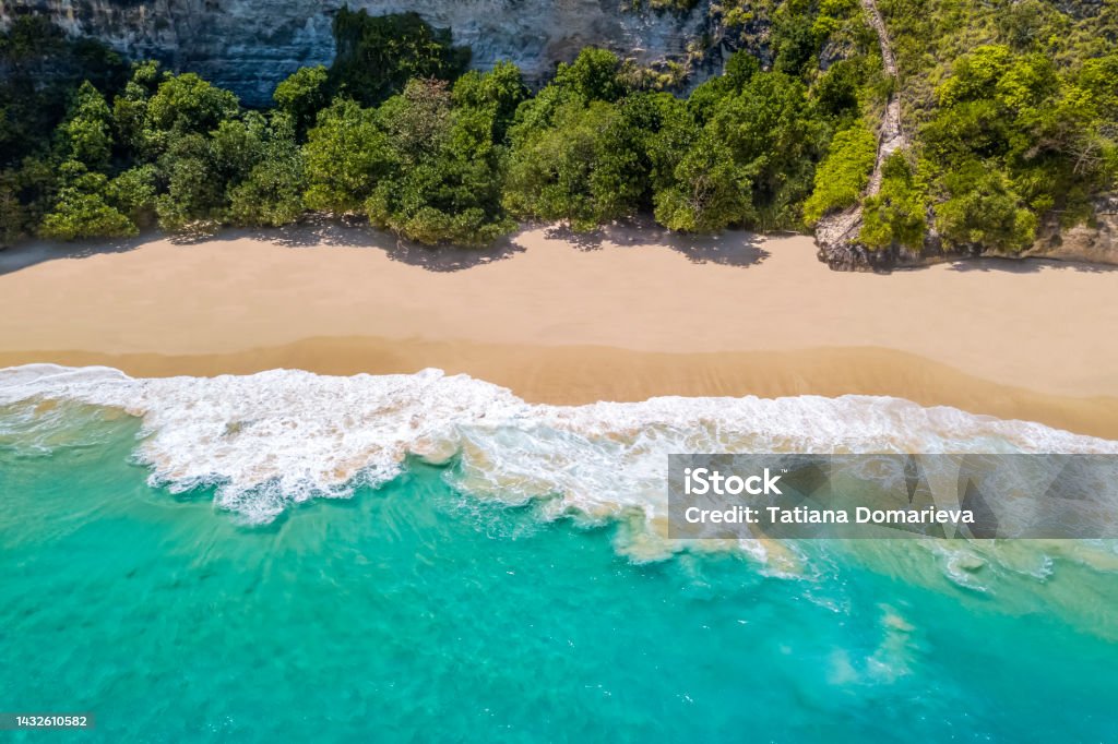 Nusa Penida Island, Kelingking Beach. Top view aerial view of a sandy clean beach with clear blue and turquoise water. Rocks and waves with white foam Nusa Penida island, Kelingking beach. Top view aerial shot of a sandy clean beach with clear blue and turquoise water. Rocks and waves with white foam Beach Stock Photo