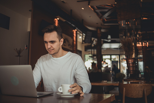 A young man sits in a cafe and works on a laptop, drinks coffee and works online in a relaxed atmosphere of the cafe