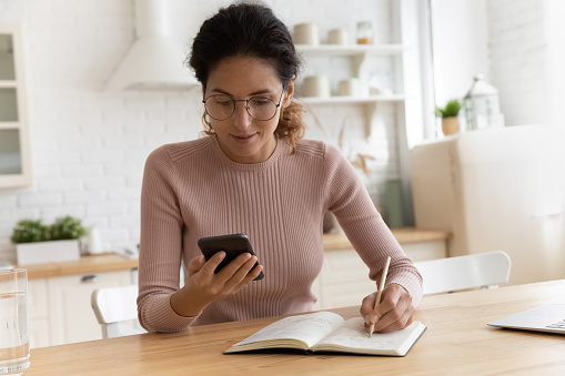 Confident woman in glasses looking at smartphone, taking notes in notebook, checking, writing down important information, focused businesswoman planning workday, meetings, student watching webinar
