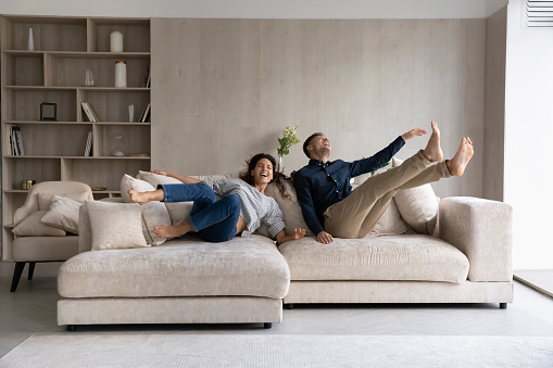 Funny excited millennial spouses of new homeowners falling on big soft couch at home. Young couple celebrating moving into first common house. Real estate, renovation, furniture for hoe concept