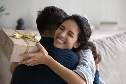 Grateful happy young woman holding festive wrap, hugging beloved husband, boyfriend, thanking for present. Sweet millennial couple celebrating anniversary, 8 march, birthday, exchanging gifts