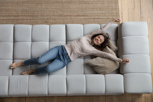 Happy peaceful young woman relaxing on comfortable couch at home with closed eyes, stretching hands and body, smiling, laughing, enjoying relaxation, leisure, weekend, lying on back. Full length