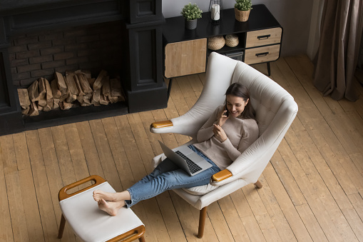 Happy young freelance employee woman talking on mobile phone at home, enjoying call, using laptop computer, relaxing in comfortable armchair in living room. Communication concept. High angle