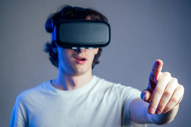 portrait of a young man in virtual reality points points his finger gray wall background. The concept of games in the mask of virtual reality.
