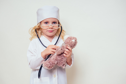 Adorable child girl uniformed wearing doctor's cap and glasses playing doctor and listening teddy bear with stethoscope on white background.kid female doctor career guidance future profession