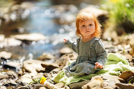 Cute little girl is sitting near the stream, space for text. Red hair baby is spending time on nature