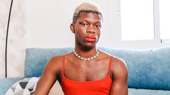 Transgender woman portrait with makeup and a red dress looking at camera while sitting on a sofa at home.