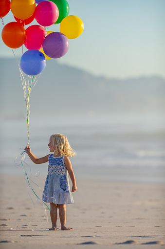 Little girl standing barefoot on the sandy beach holding a bunch of colorful balloons