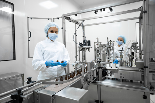 Two employees in pharmaceutical industry wearing protective gloves, mask, cap and white suit seen standing by the machine that is the part of the medicaments production during the working hours in a pharmaceutical manufacturing.