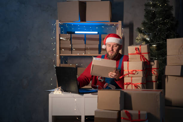 산타 클로스는 크리스마스 이브 늦게 홈 오피스 테이블에 앉아있는 노트북 컴퓨터를 사용하여 선물 상자를 들고 모자를 쓰고 있습니다. 전자 상거래 웹 사이트 크리스마스 휴일 온라인 쇼핑 전� - santa claus christmas watch workshop 뉴스 사진 이미지