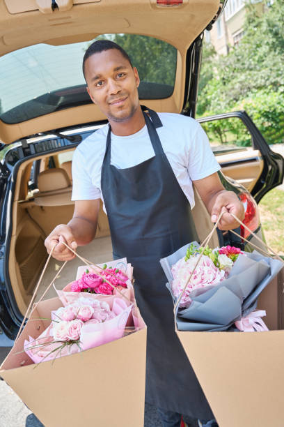 jovem com flores em sacos fica na frente de um carro - men african descent giving flower - fotografias e filmes do acervo