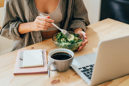 Unrecognizable woman eating salad at workplace in office in front of laptop.