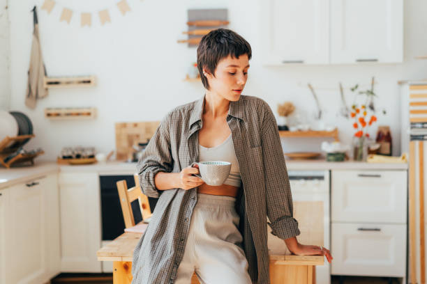 eine junge frau mit kurzen haaren im pyjama und hemd trinkt kaffee in der küche. - stereotypical housewife women domestic kitchen brown hair stock-fotos und bilder