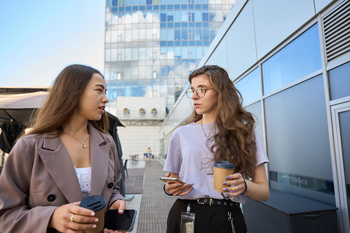Focused woman walking down street near office next to colleague while they are drinking coffee and holding smartphones