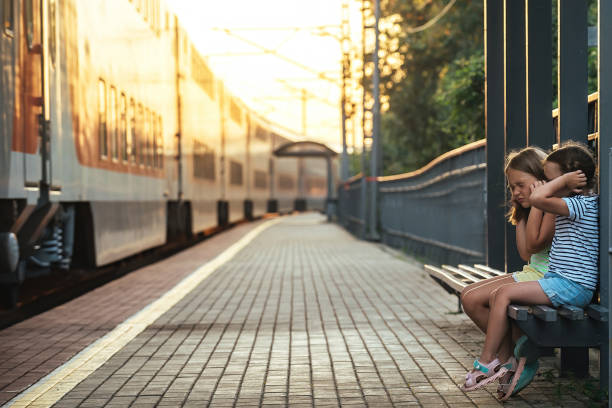two little girls are sitting on a bench of the railway station and, out of fear of a loud sound passing by a train, they covered their ears with their hands so as not to hear