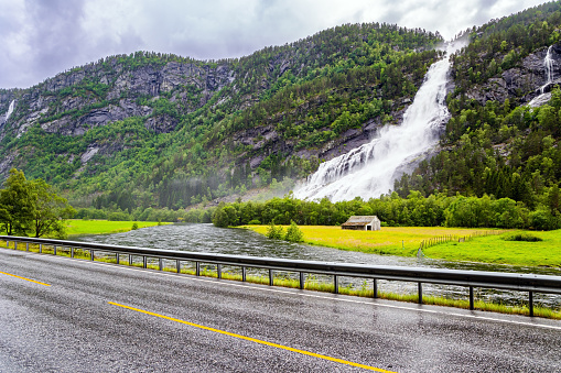 Huge powerful cascading waterfal Vidfossen. The rain-wet highway glitters in the fog. Western Norway. White water foam envelops the foot of the waterfall. Summer