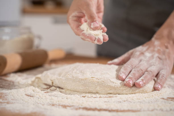 istock Women's hands pouring flour into raw dough while kneading bread dough on a table full of flour in the bakery kitchen. 1432694087