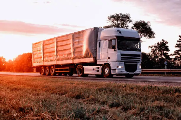 Photo of A truck with a tilt semi-trailer transports cargo against the backdrop of a sunset in the summer. Sharing economy concept in freight logistics. Modernization of logistics companies