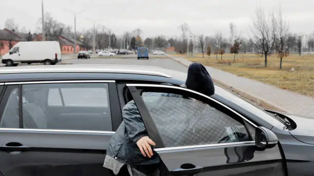 Photo of Homeless thief steals purse items money from car in public parking. Auto with unlocked doors open. Property insurance against unlawful theft or crime. Male burglar looks through crack in window.