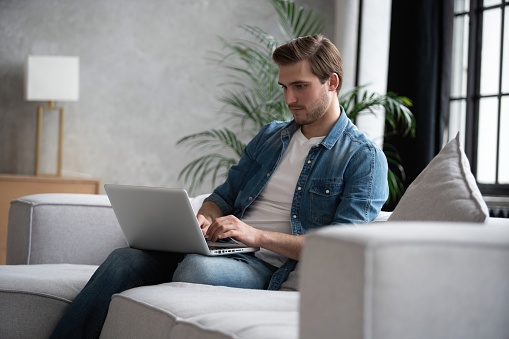 Young attractive smiling man is browsing at his laptop, sitting at home on the cozy sofa at home, wearing casual outfit