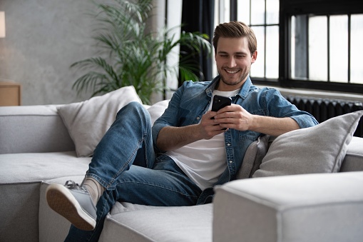 Portrait of an attractive smiling young man wearing casual clothes sitting on a couch at the living room, using mobile phone