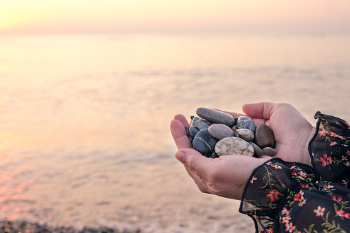 Women's hands hold large pebbles in their palms against the backdrop of calm sea water in the light of the setting sun. The concept of sea holidays, a romantic vacation on the coast.
