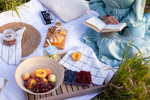 Cropped photo of woman in long dress with short hair sitting on a white blanket with fruits and pastries and reading the book. Concept of having picnic in a city park during summer holidays or weekend