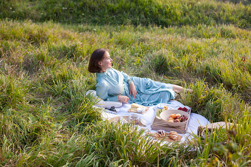 A girl in a long summer dress with short hair lying on a white blanket with fruits and pastries and enjoying the view. Concept of having picnic in a city park during summer holidays or weekends.