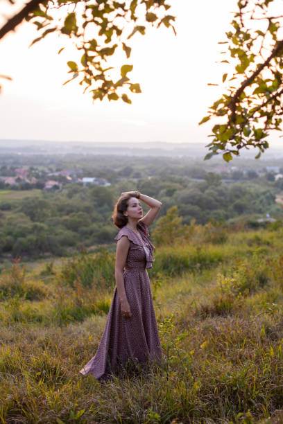 Beautiful young smiling girl in a long brown dress stands along the lawn. Happy woman walks at sunset on a hill overlooking the river. Concept of having rest in park during summer holidays or weekends
