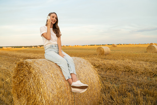 Attractive stylish charming girl with a happy smile talking on the phone sitting on a hay stack in a field.