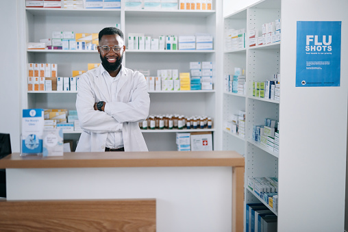 Portrait of black man pharmacist in a pharmacy with medicine, pills and flu treatment. Healthcare worker standing behind the counter, smiling and ready to serve medication and prescription in clinic