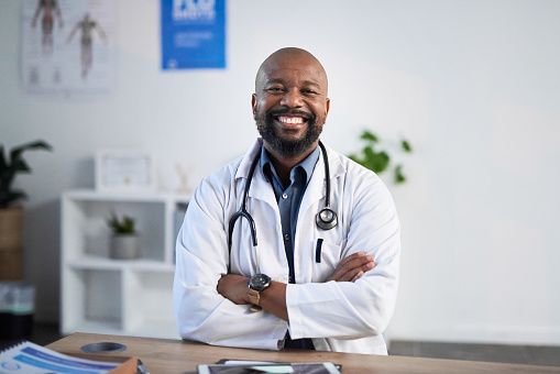 Medical, portrait and African doctor consulting with a smile at a table in his office at a hospital. Black man, surgeon or cardiology worker with pride and arms crossed while working at a clinic