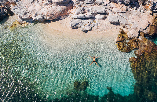 Aerial photo of man lying on the water in STAR pose on rocky pebbly beach and sun tanning. Soft waves washing his body on lonely deserted Greek island in Ionian Sea . Exotic vacation concept