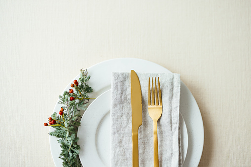 Flat lay of a holiday table setting with golden cutlery on a napkin on top of a white porcelain plat with a small conifer branch on the side.