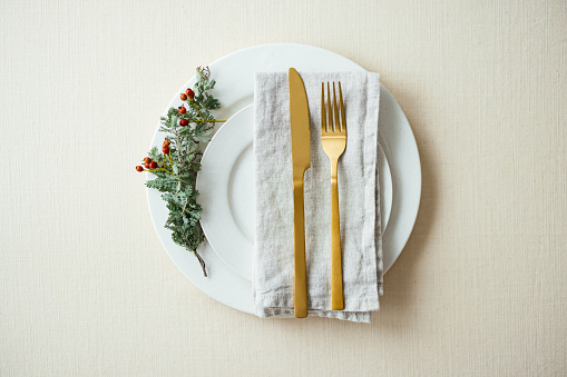 Flat lay of a holiday table setting with golden cutlery on a napkin on top of a white porcelain plat with a small conifer branch on the side.