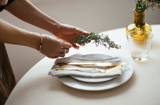 Close up shot of an unrecognizable woman's hands setting up holiday table at home. She is putting golden cutlery and porcelain plate on the table and decorating it with a small branch on the side.