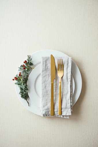 Flat lay of a holiday table setting with golden cutlery on a napkin on top of a white porcelain plat with a small conifer branch on the side.