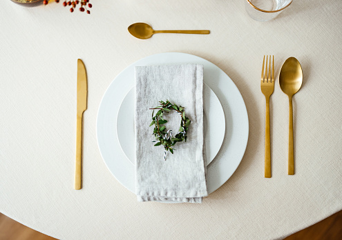 Elegant table setting with golden cutlery, porcelain plates and a napkin with olive branch decoration on top.